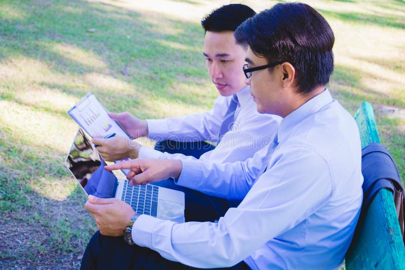 Businessman,They are sitting on bench in park.He is play notebook and  search internet.Another one holding a cup of coffee and talking about business in  relax time.Photo concept  business