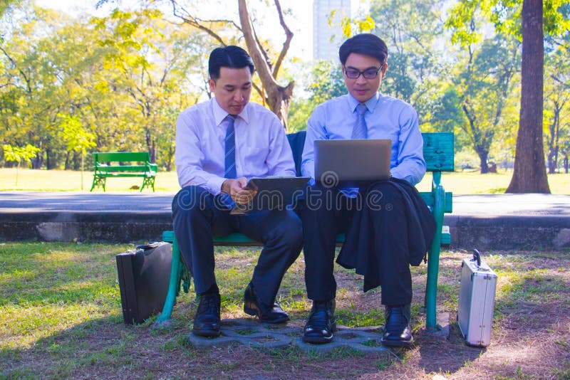 Businessman,They are sitting on bench in park.He is play notebook and  search internet.Another one holding a cup of coffee and talking about business in  relax time.Photo concept  business