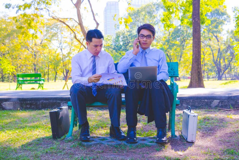 Businessman,They are sitting on bench in park.He is play notebook and  search internet.Another one holding a cup of coffee and talking about business in  relax time.Photo concept  business