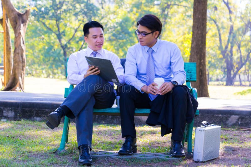 Businessman,They are sitting on bench in park.He is play notebook and  search internet.Another one holding a cup of coffee and talking about business in  relax time.Photo concept  business