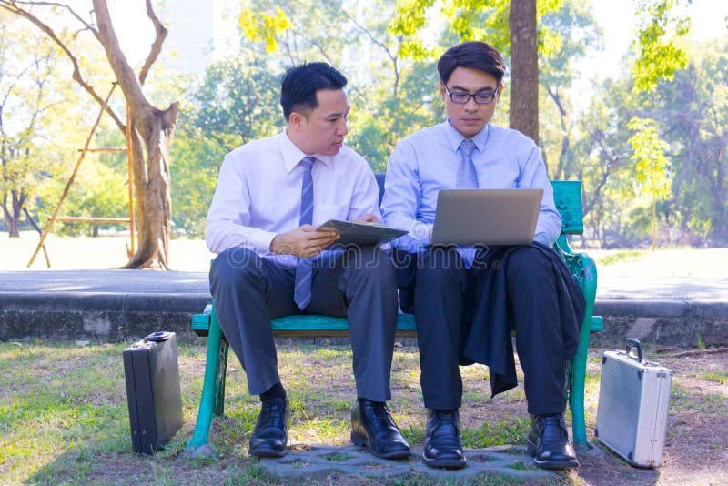 Businessman,They are sitting on bench in park.He is play notebook and  search internet.Another one holding a cup of coffee and talking about business in  relax time.Photo concept  business