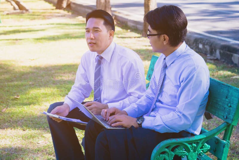 Businessman,They are sitting on bench in park.He is play notebook and  search internet.Another one holding a cup of coffee and talking about business in  relax time.Photo concept  business