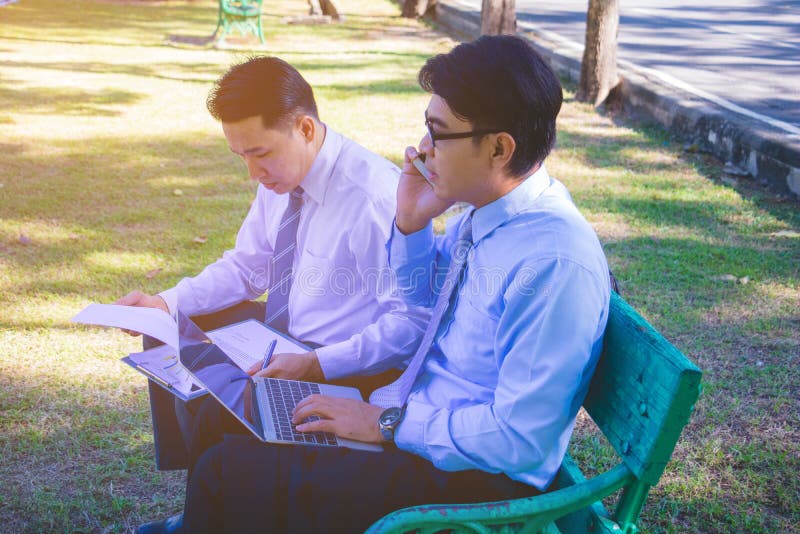 Businessman,They are sitting on bench in park.He is play notebook and  search internet.Another one holding a cup of coffee and talking about business in  relax time.Photo concept  business