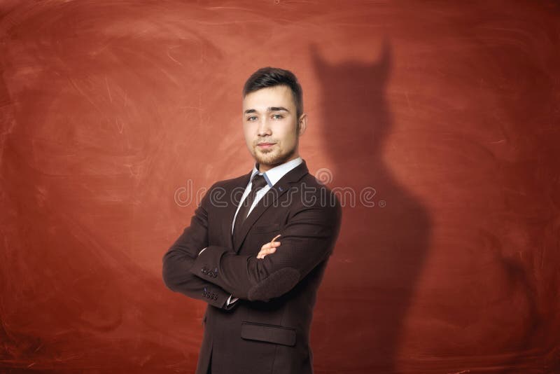 Businessman in suit standing with his arms folded, he is casting shadow of the devil on the rusty orange wall behind him stock images