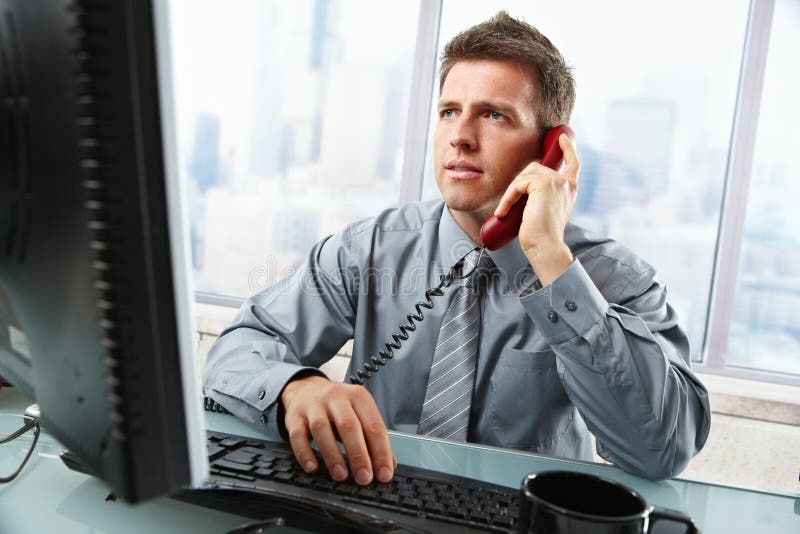 Determinato imprenditore discutere il lavoro al computer a telefono fisso, mentre guardando la schermata di digitazione su tastiera in ufficio.