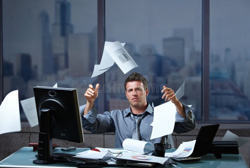 Exhausted businessman throwing documents into air sitting at office desk in overtime.