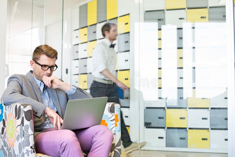 Businessman Using Laptop On Armchair With Colleague Walking In