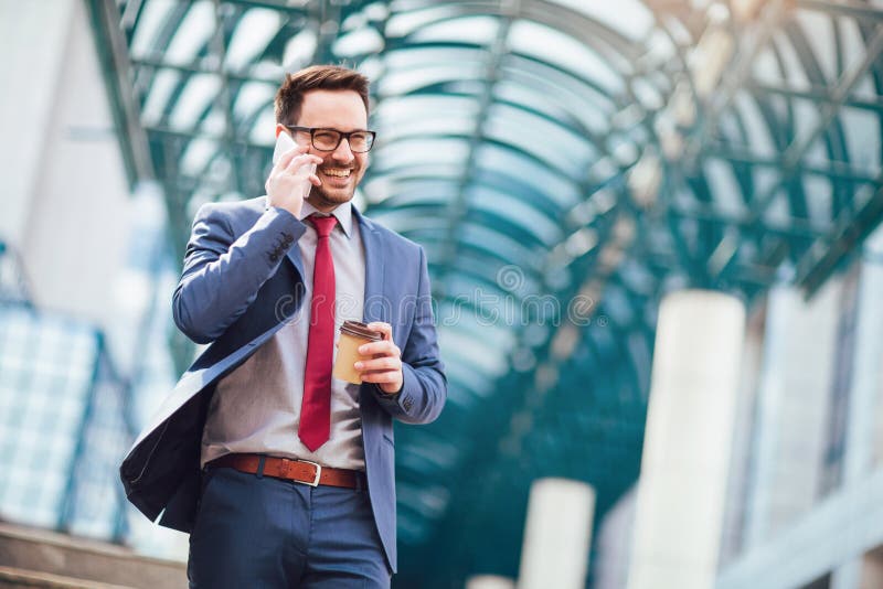 Businessman using mobile phone outside of office buildings in the background. Young caucasian man holding smartphone for business work and drink coffee to go.