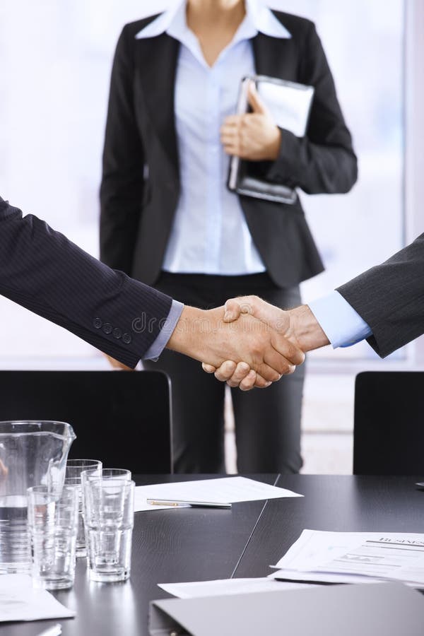 Businessmen shaking hands in office, assistant in background, handshake in closeup.