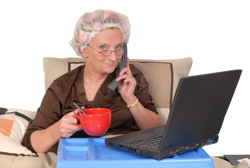 Middle aged businesswoman in pyjamas working at home in bed, laptop on tray. Making phone call. Middle aged businesswoman in pyjamas working at home in bed, laptop on tray. Making phone call