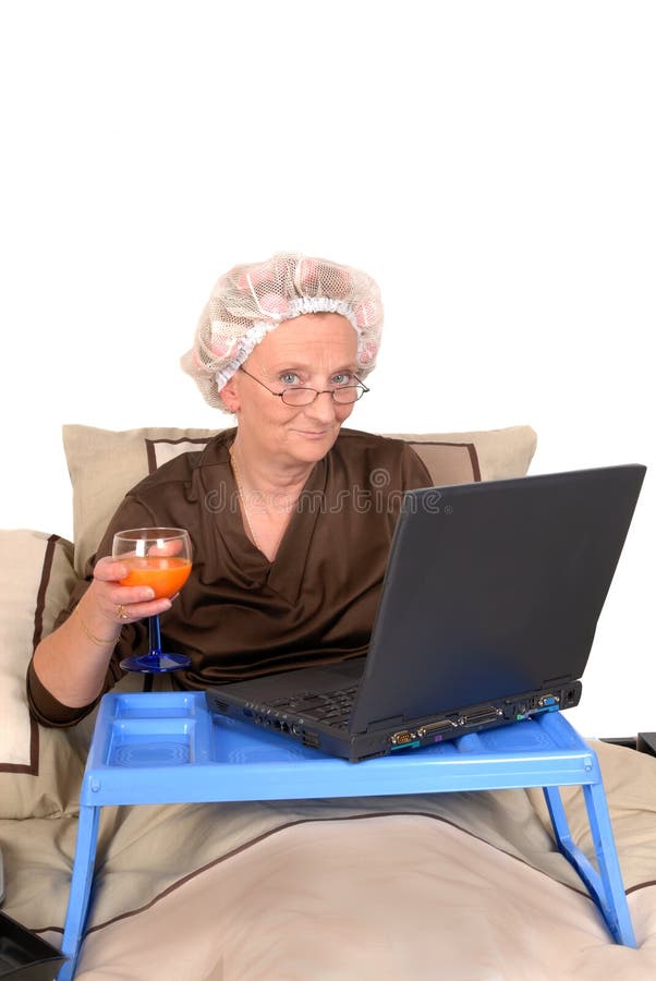 Middle aged businesswoman in pyjamas working at home in bed, laptop on tray. Curlers and net in hair. Middle aged businesswoman in pyjamas working at home in bed, laptop on tray. Curlers and net in hair.