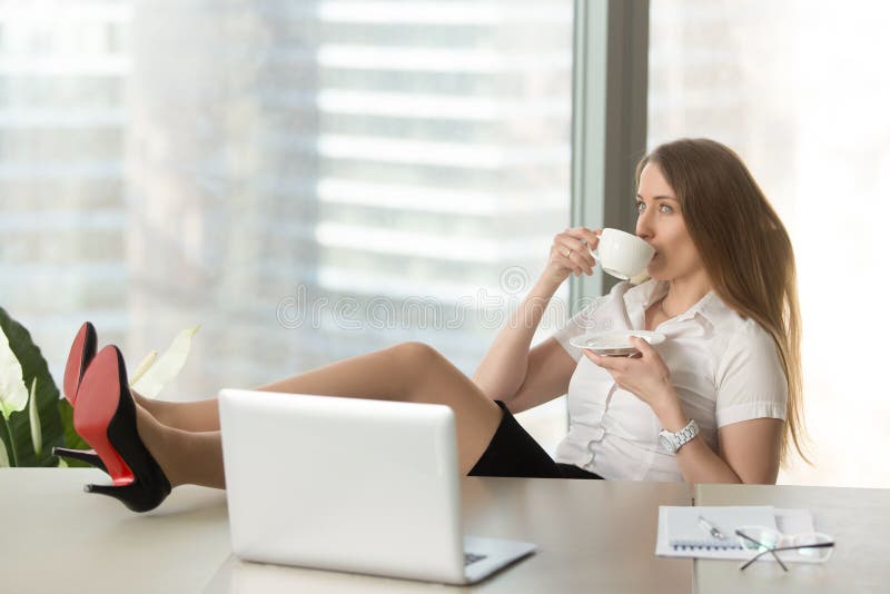 Businesswoman drinking coffee with legs on desk. Girl resting at the desk with cup of tea. Confident lady takes a break at work. Successful female entrepreneur relaxing after productive day in office