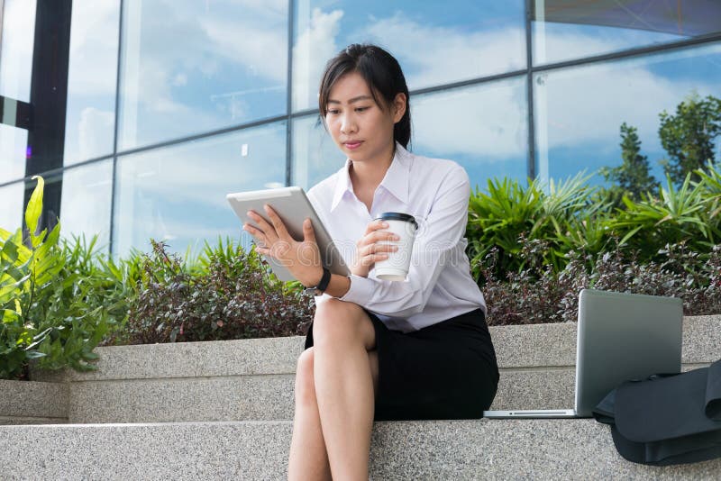 businesswoman holding digital tablet & coffee outside office building. beautiful young asian woman with tea sitting outdoors. fem royalty free stock photos businesswoman holding digital tablet & coffee outside office building. beautiful young asian woman with tea sitting outdoors. fem royalty free stock photos