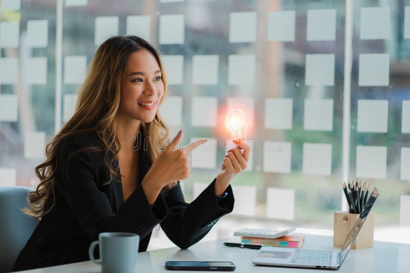 A businesswoman in a suit holds a light bulb and uses a laptop on a wooden table. Creativity with innovation and inspiration save