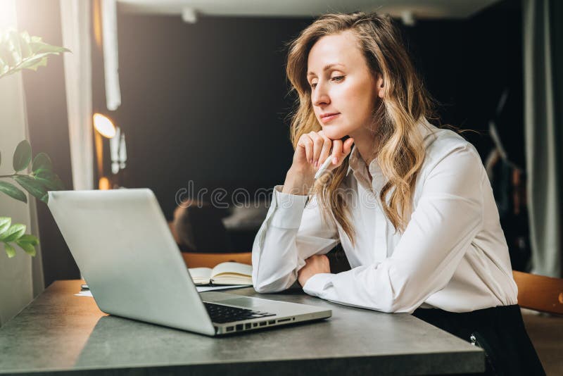 Young businesswoman in white shirt is sitting in office at table in front of computer and pensively looks at screen of laptop, holding pen in her hand. Fatigue from work, sadness. Distance education.