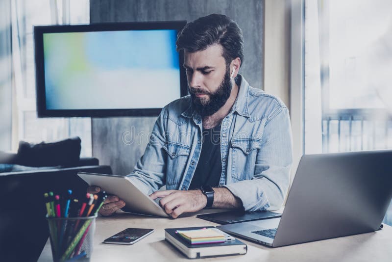 Young man sitting at window and working on laptop. Bearded man sitting at  desktop with smartphone in his hands. Online education. Stock Photo