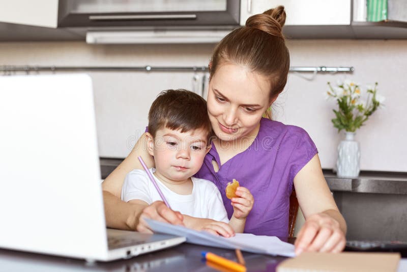 Busy working mother sits in front of opened laptop computer, tries to conecntrate on work, sits against kitchen interior with chil stock image Busy working mother sits in front of opened laptop computer, tries to conecntrate on work, sits against kitchen interior with chil stock image