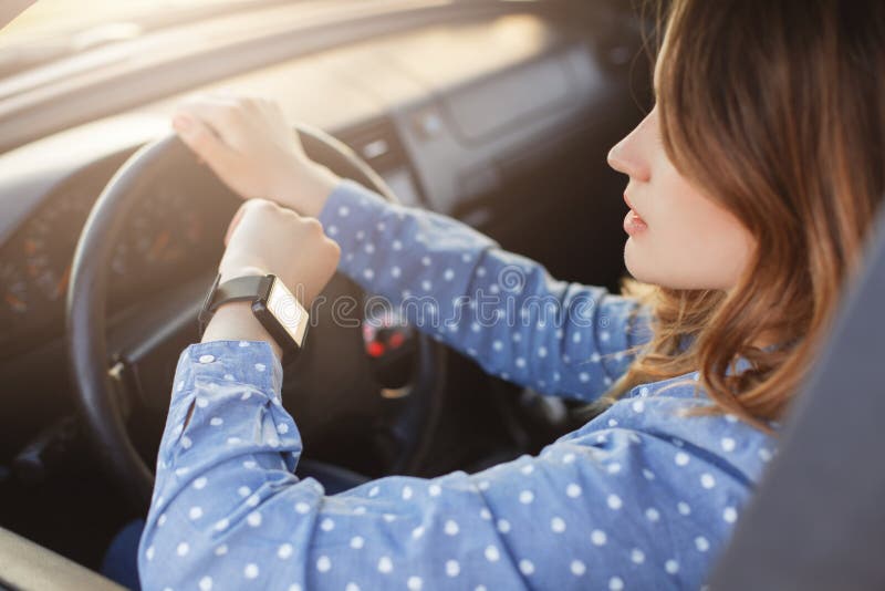 Busy young woman drives car and looks at watch, stuck in traffic jam, hurries to work, being nervous and stressed, feels impatient