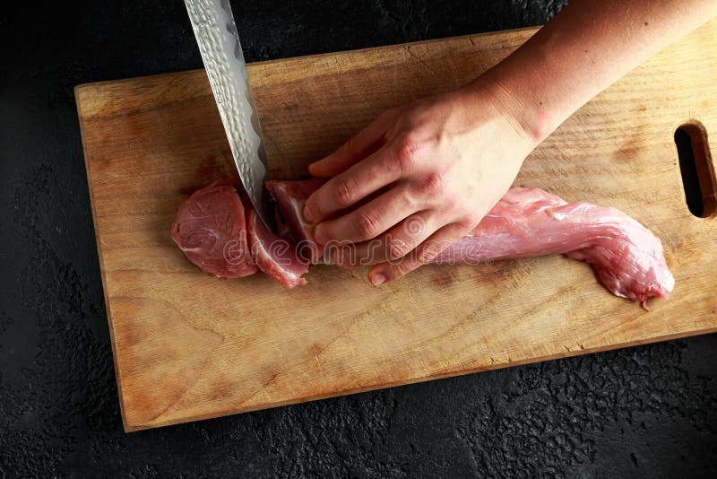 Butcher`s hands ready to cut with a knife raw pork tenderloin fillet on wooden board.