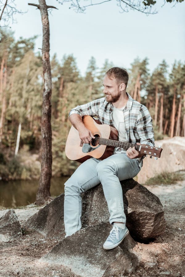 Bärande jeans för stilig skäggig man och kvadrerad skjorta som spelar gitarren nära sjön royaltyfri fotografi Bärande jeans för stilig skäggig man och kvadrerad skjorta som spelar gitarren nära sjön royaltyfri fotografi