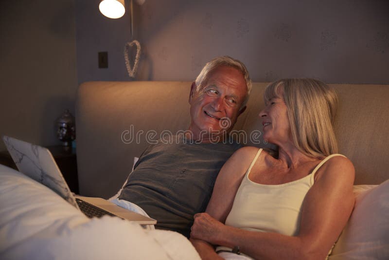 Bärande pyjamas för höga par som ligger i säng som ser bärbara datorn royaltyfri foto Bärande pyjamas för höga par som ligger i säng som ser bärbara datorn royaltyfri foto