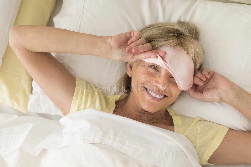 High angle portrait of happy mature woman wearing sleep mask while lying on bed. High angle portrait of happy mature woman wearing sleep mask while lying on bed