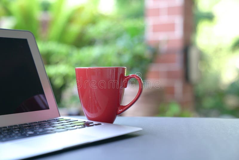 Bärbar dator och kaffe i rött koppel med trädgårdbakgrund. arbete hemma fotografering för bildbyråer