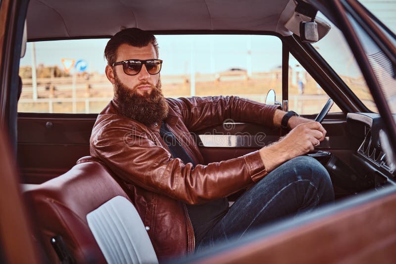 Country style concept. Bearded male in sunglasses dressed in brown leather jacket driving a retro car. Country style concept. Bearded male in sunglasses dressed in brown leather jacket driving a retro car.