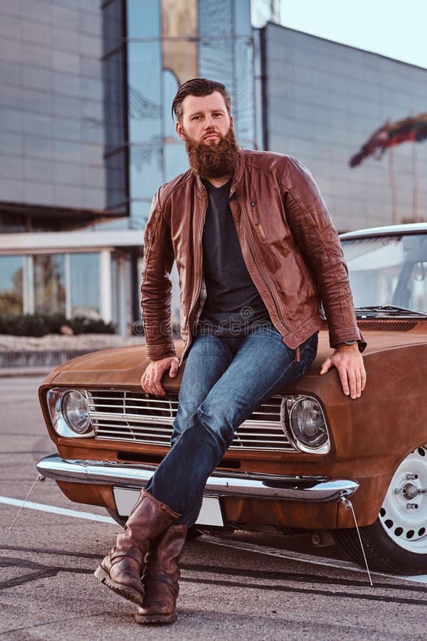 Country style concept. Bearded male dressed in brown leather jacket and boots leaning on tuned retro car in the city parking near skyscraper. Country style concept. Bearded male dressed in brown leather jacket and boots leaning on tuned retro car in the city parking near skyscraper.