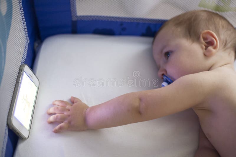 Bébé bandes dessinées de observation d'une berceuse avec le téléphone portable sur la huche photo stock Bébé bandes dessinées de observation d'une berceuse avec le téléphone portable sur la huche photo stock