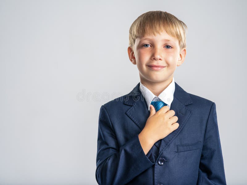 Băiat blond pozitiv îmbrăcat într-un costum formal atingându-și cravata. portretul unui copil alb zâmbitor într-un costum albastru stock photography