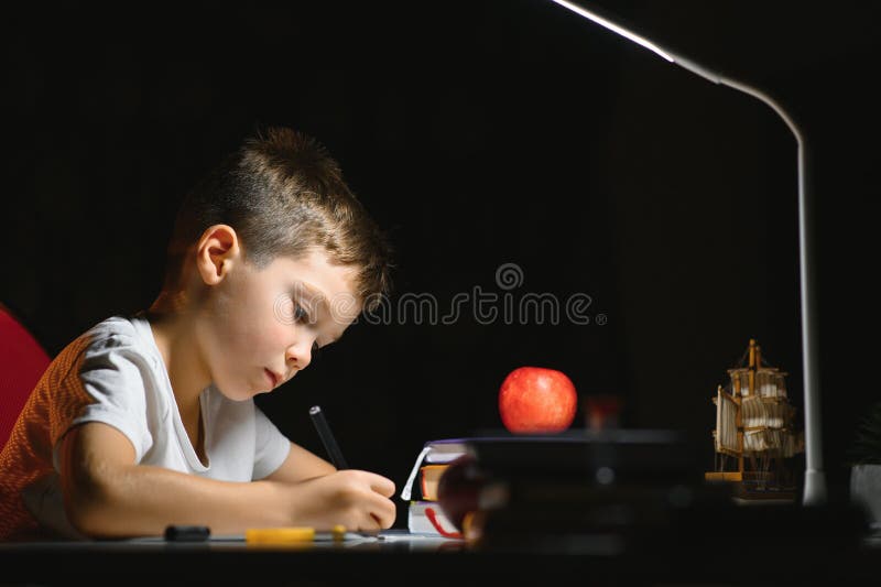 Băiatul învață lecții de acasă la masă în lumina unei lămpi de masă royalty free stock images