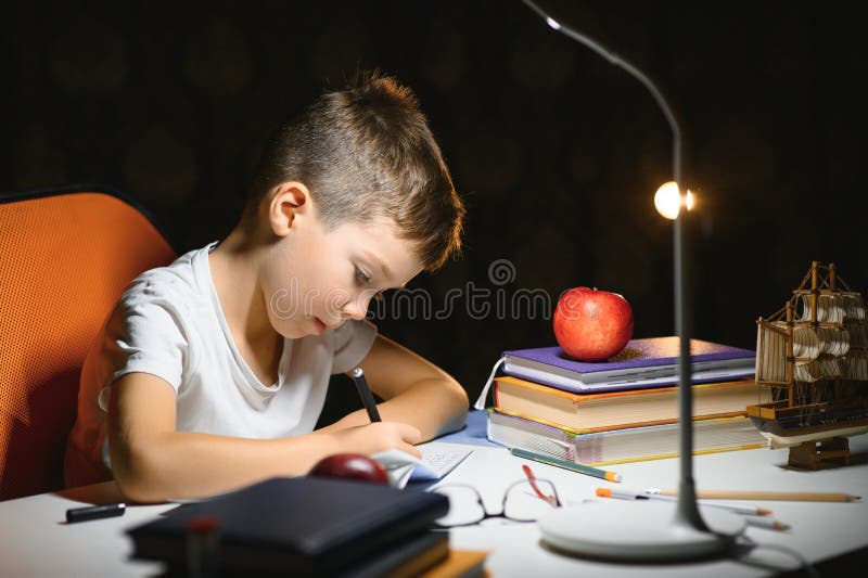 Băiatul învață lecții de acasă la masă în lumina unei lămpi de masă stock photos