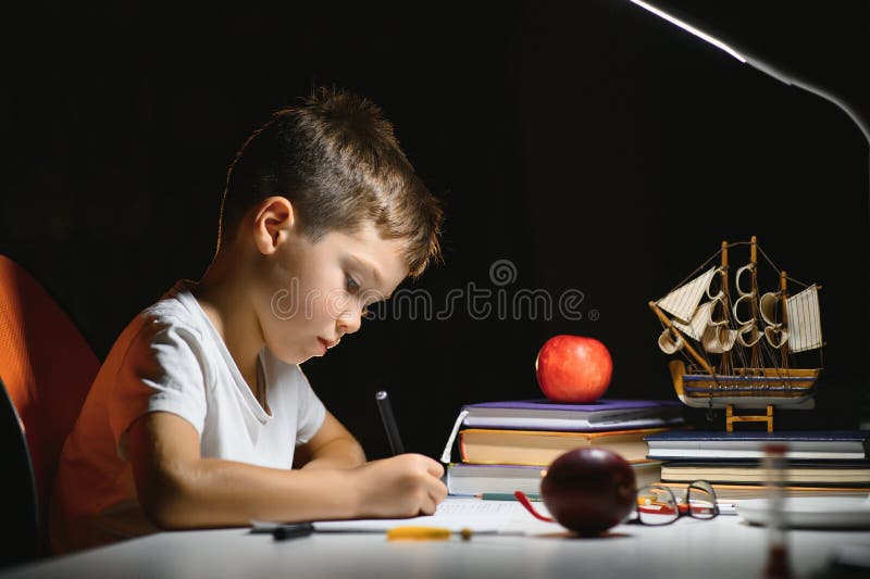 Băiatul învață lecții de acasă la masă în lumina unei lămpi de masă stock photos