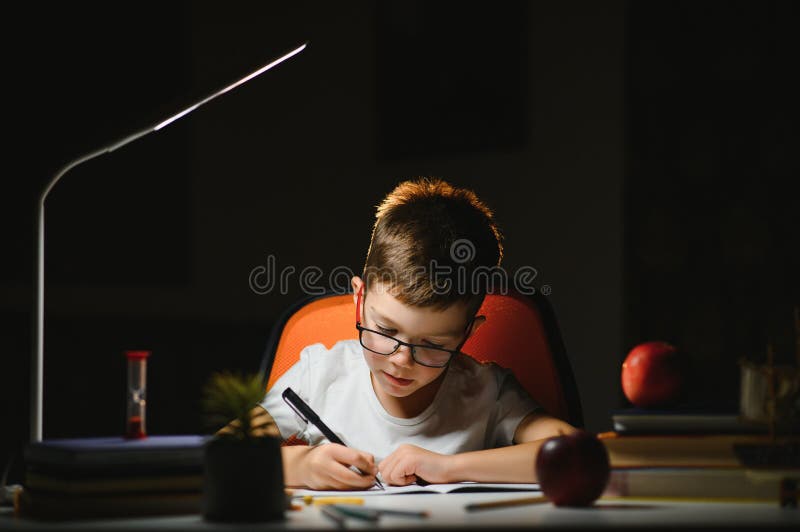Băiatul învață lecții de acasă la masă în lumina unei lămpi de masă stock photo