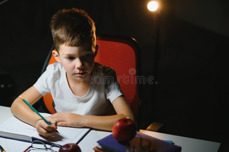 Băiatul învață lecții de acasă la masă în lumina unei lămpi de masă royalty free stock images