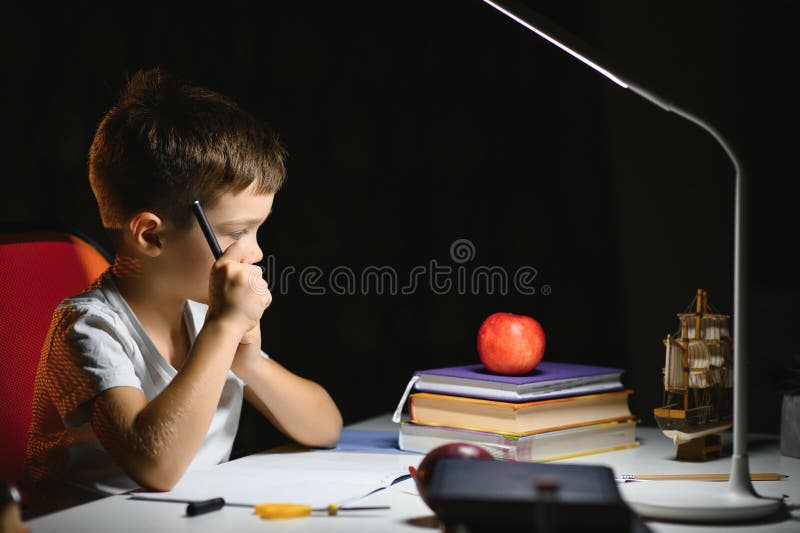 Băiatul învață lecții de acasă la masă în lumina unei lămpi de masă stock images