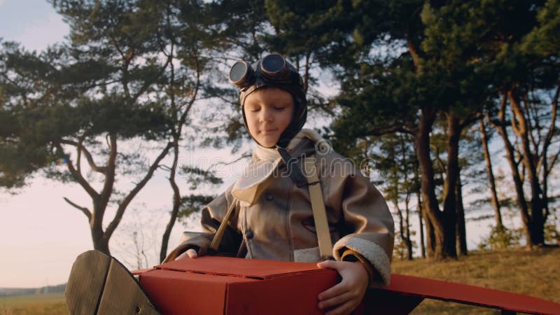 Băiețel pilot pozitiv sub arbori în avion cardboard distractiv costum poartă ochelari retro-aviator super-erou cu mișcare lentă stock video footage