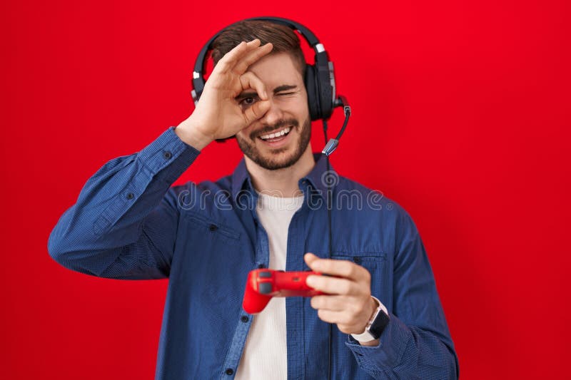 Bărbat hispanici cu barbă jucând jocuri video, cu controlerul zâmbind fericit făcând semn cu mâna în ochi uitându-se printre deget stock photos