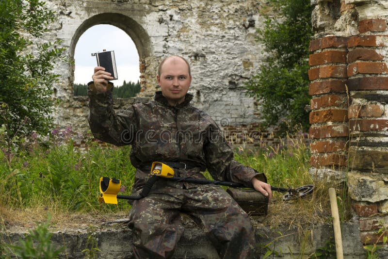 Man treasure hunter sitting in the ruins of a building with a metal detector on his knees, solemnly raises a flask, holding his second hand on old chest. Man treasure hunter sitting in the ruins of a building with a metal detector on his knees, solemnly raises a flask, holding his second hand on old chest