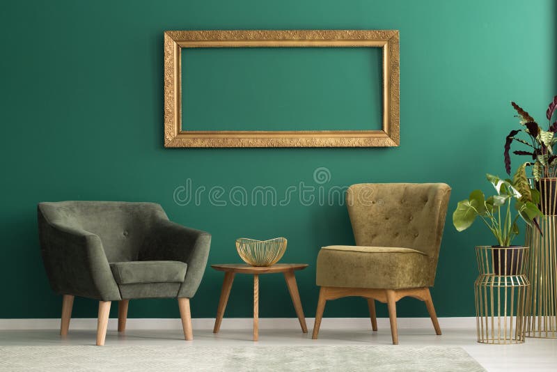 Empty frame above retro, upholstered chairs in a green living room interior with plants and golden decorations. Empty frame above retro, upholstered chairs in a green living room interior with plants and golden decorations