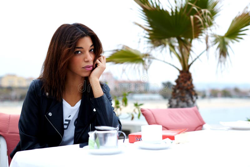 Charming fashionable female posing to the camera while sitting in modern sidewalk cafe terrace, afro american woman drink coffee and enjoying her recreation time at weekend. Charming fashionable female posing to the camera while sitting in modern sidewalk cafe terrace, afro american woman drink coffee and enjoying her recreation time at weekend