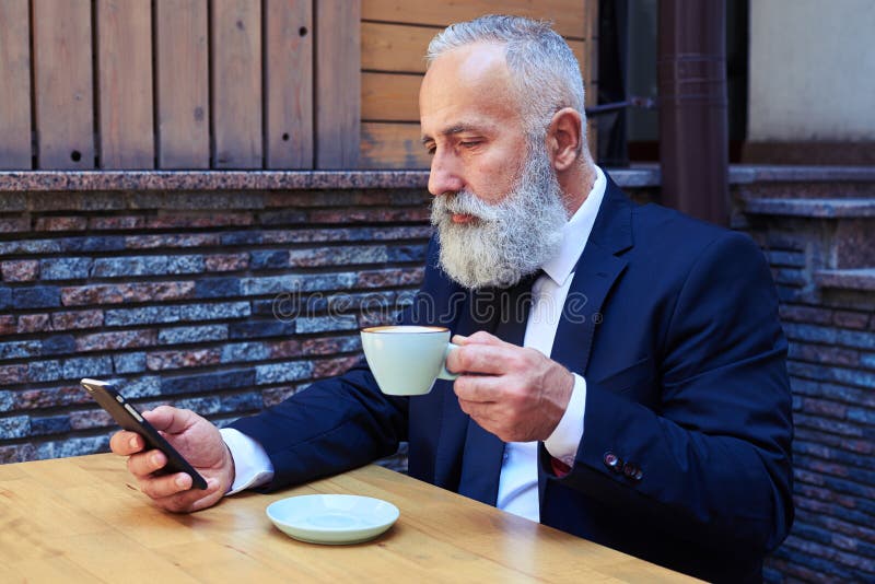 Caffè bevente dell'uomo anziano bello mentre praticando il surfing in telefono cellulare fotografia stock libera da diritti Caffè bevente dell'uomo anziano bello mentre praticando il surfing in telefono cellulare fotografia stock libera da diritti