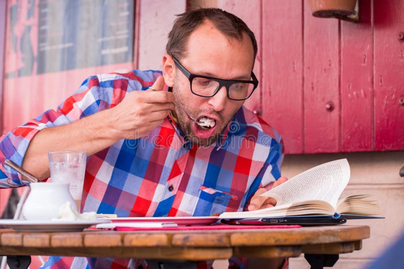 Café bebendo do homem considerável novo em um café dentro e comendo o sanduíche. Golding um livro. imagem de stock Café bebendo do homem considerável novo em um café dentro e comendo o sanduíche. Golding um livro. imagem de stock