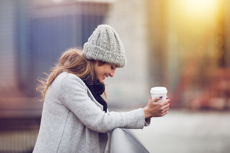 Beautiful happy young adult woman drinking coffee near New York City skyline wearing winter clothes and smiling. Beautiful woman holding coffee cup outdoors in the city. Beautiful happy young adult woman drinking coffee near New York City skyline wearing winter clothes and smiling. Beautiful woman holding coffee cup outdoors in the city.