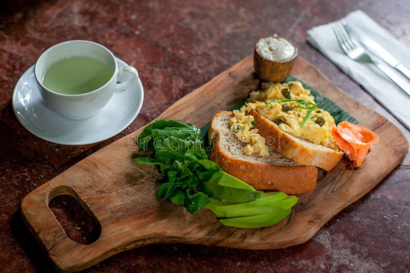 Café da manhã saudável com brinde e ovos mexidos do pão de Wholemeal com avokado, queijo e salmões na placa de corte e no chá ver fotografia de stock Café da manhã saudável com brinde e ovos mexidos do pão de Wholemeal com avokado, queijo e salmões na placa de corte e no chá ver fotografia de stock