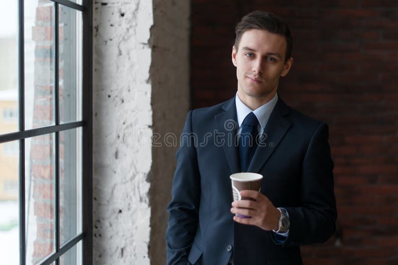 Café de consumición del hombre de negocios en la oficina que coloca la ventana cercana que mira la cámara imagen de archivo Café de consumición del hombre de negocios en la oficina que coloca la ventana cercana que mira la cámara imagen de archivo