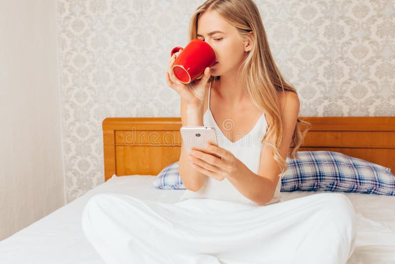 Café de consumición de la muchacha hermosa joven, sentándose en cama y la mirada foto de archivo libre de regalías Café de consumición de la muchacha hermosa joven, sentándose en cama y la mirada foto de archivo libre de regalías