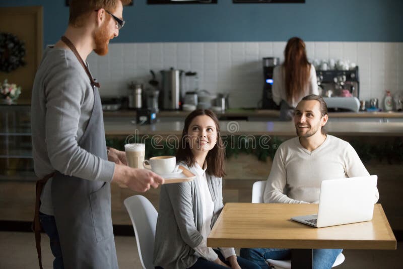 Café de transport de serveur aux couples de sourire attendant à la table de café image stock Café de transport de serveur aux couples de sourire attendant à la table de café image stock