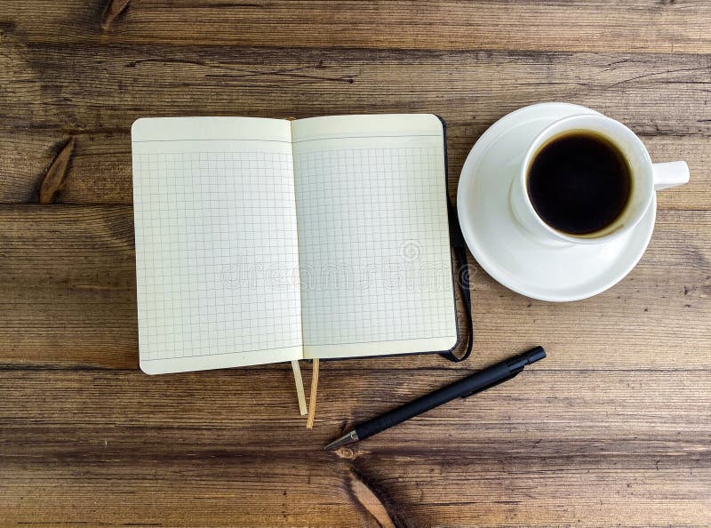 Coffee in a cup and a notebook with a pen on the table, top view. Coffee in a cup with a saucer and a notebook with a pen on a wooden table. Coffee in a cup and a notebook with a pen on the table, top view. Coffee in a cup with a saucer and a notebook with a pen on a wooden table